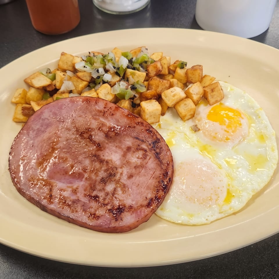 A breakfast plate with a grilled ham steak, two fried eggs, and home fries topped with onions and peppers.