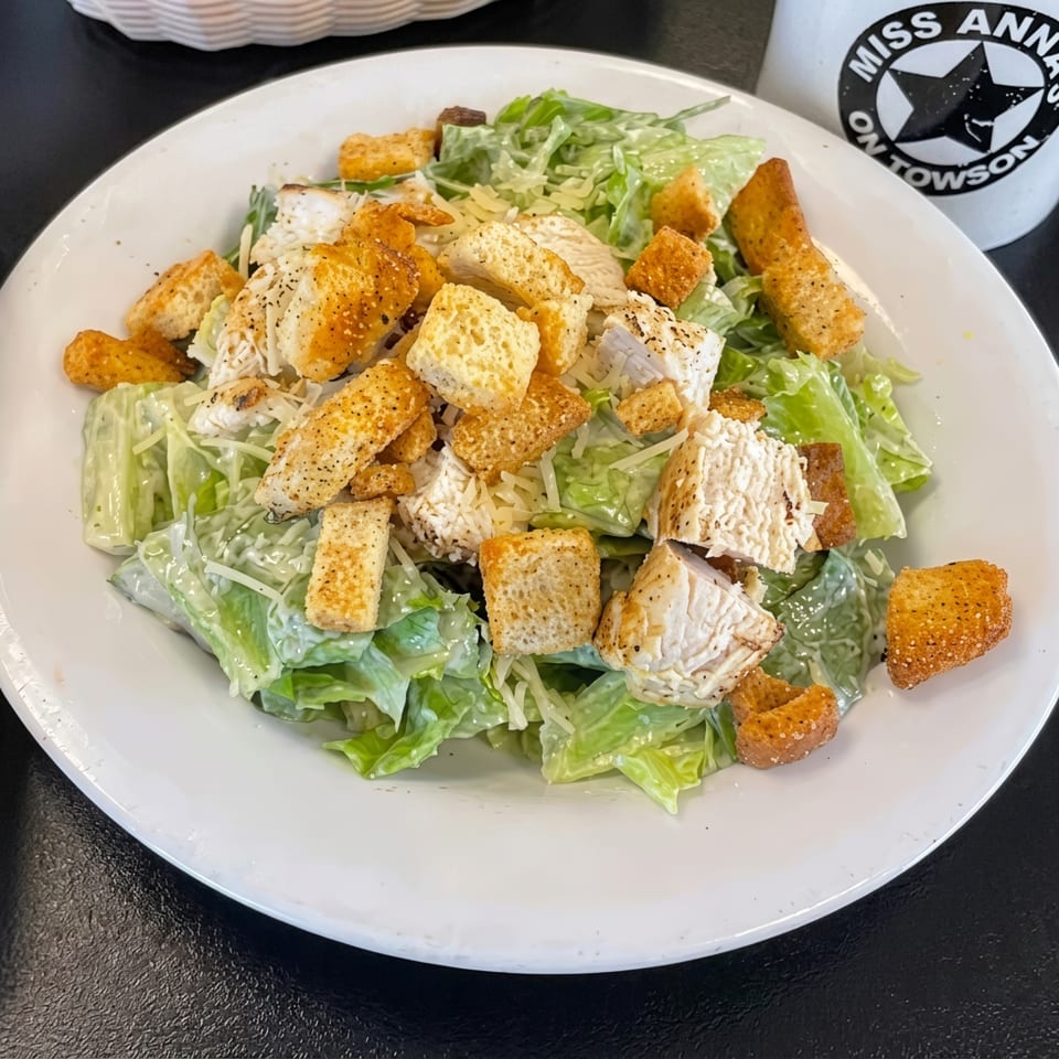 A Caesar salad with chicken, croutons, and shredded cheese on a white plate, with a Miss Anna's mug.
