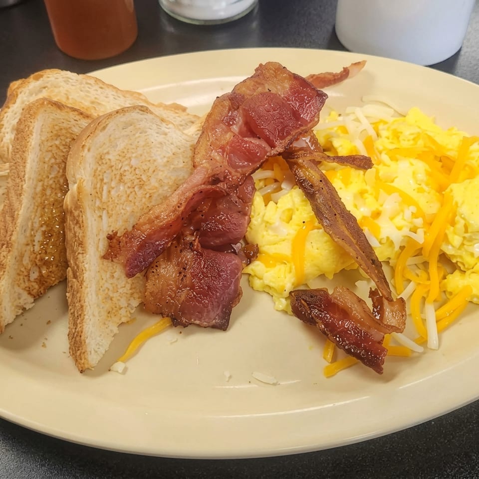 A breakfast plate with crispy bacon, scrambled eggs with cheese, and three slices of toast.