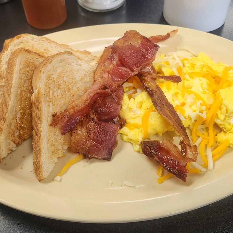 A breakfast plate with crispy bacon, scrambled eggs with cheese, and three slices of toast.
