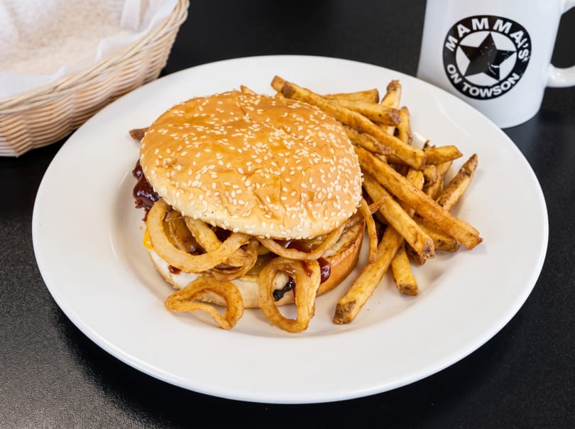 A burger with onion rings and fries on a white plate, with a "Mamma's on Towson" mug.
