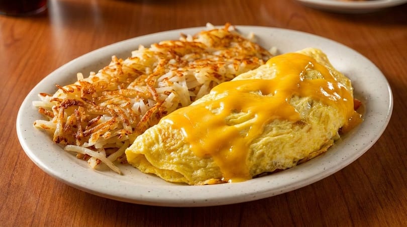A cheesy omelet and crispy hash browns are served on a white plate on a wooden table.